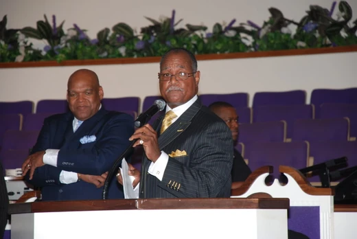 A speaker with a microphone at the church pulpit, with people seated behind him in the sanctuary.